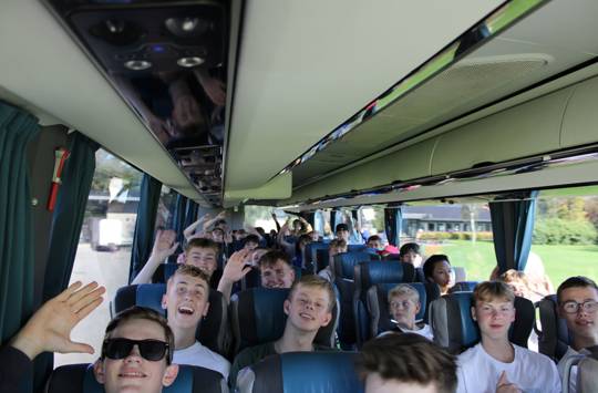 En flok glade efterskoleelever sidder i en bus og smiler og vinker. 