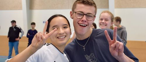 To efterskoleelever står smilende i en hal og giver tegn på sejr med fingrene